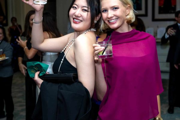 Two women smiling at a social event, holding drinks, dressed elegantly.