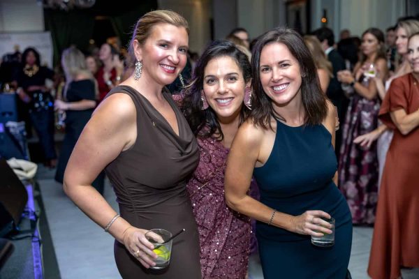 Three women smiling at the Believe Ball event, holding drinks, in a lively social setting.