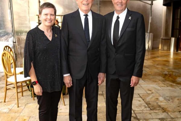 Three smiling people in formal attire at Believe Ball event under a tent.