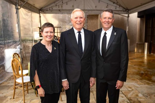 Three smiling people in formal attire at Believe Ball event in a tent.