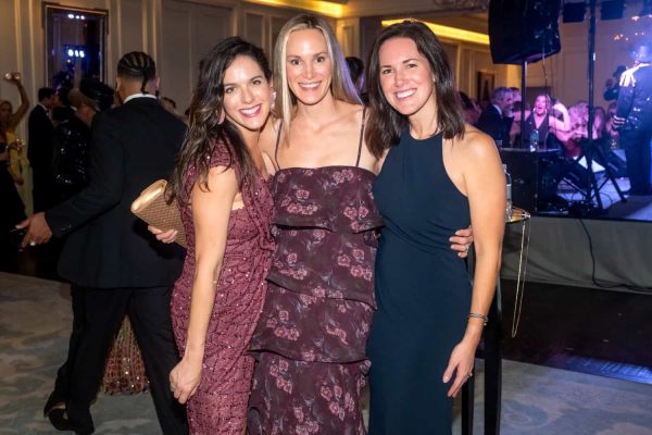 Three women smiling at the Believe Ball event in formal attire.