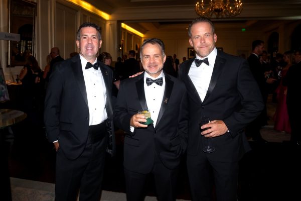 Three men in tuxedos at a formal event, smiling and holding drinks.