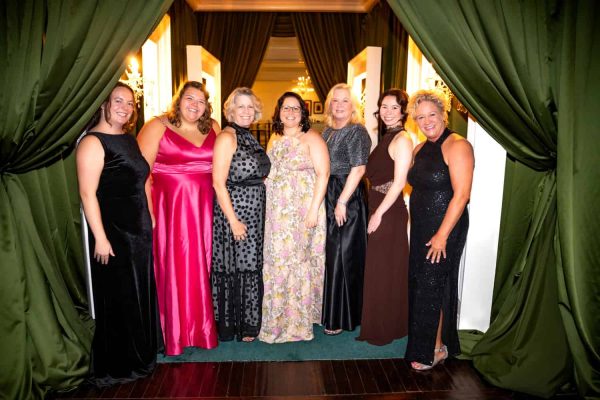 Group of women in elegant dresses at Believe Ball event in front of green curtains.