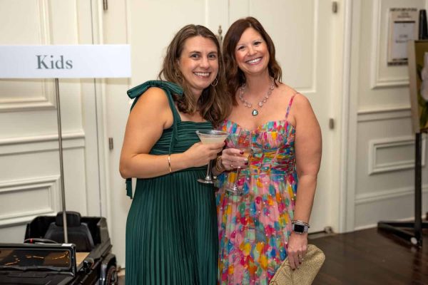 Two women smiling at a Believe Ball event, holding drinks, in colorful dresses.
